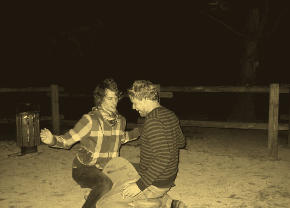 Two young men crouched on a small playground seesaw at night, laughing.